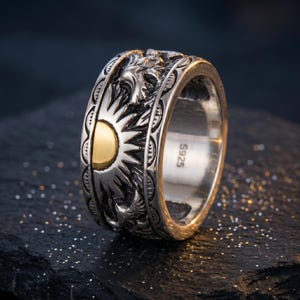 May include: A wide silver band ring featuring an embossed sun motif with a polished gold-tone center. The exterior surface displays intricate carved details and patterns surrounding the sun. The interior of the band is stamped with S925.