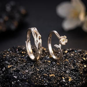 Könnte beinhalten: Zwei silberne Ringe mit naturinspirierten Designs. Ein Ring hat ein strukturiertes, astähnliches Muster mit offenen Aussparungen. Der zweite Ring ist ein zartes Band, das mit einer kleinen, fünfblättrigen Blume mit goldfarbener Mitte verziert ist.
