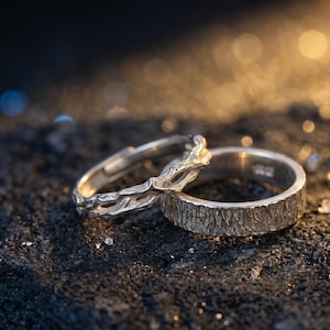 Puede incluir: Dos anillos de plata. Uno presenta un diseño texturizado similar a la corteza, mientras que el otro tiene un intrincado patrón de vid calada. Los anillos están sobre una superficie oscura y texturizada con un fondo borroso.