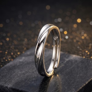 Peut inclure: Une bague réglable argentée avec un motif torsadé. La bague est posée sur une surface sombre et texturée avec un arrière-plan flou de lumières dorées et blanches. La surface de la bague reflète la lumière.