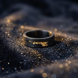 Può includere: Un anello grigio scuro con una finitura opaca e testurizzata, caratterizzato da un inserto orizzontale e irregolare in foglia d'oro brillante. La fascia ha un profilo largo e piatto con un netto contrasto tra l'esterno scuro e ruvido.