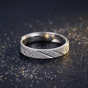 May include: A silver ring with a textured band and small, clear gemstones. The ring features a floral design and diagonal lines, set against a dark, glittery background. The ring is a piece of jewelry.