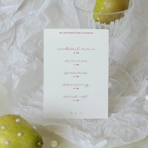 May include: A white rectangular wedding schedule card with dark red text reading AN UNFORGETTABLE EVENING, cocktail hour 5 PM, dinner 7 PM, speeches 8 PM, dancing 9 PM, send-off 11 PM, and R & A at the bottom, positioned near a glass bowl and pears.