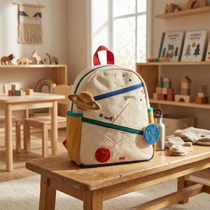 May include: A beige backpack with a red handle and colorful planet and star appliqués. The backpack has a blue and green stripe design, with the words "All True" and "Mars" on the front. A fun accessory for kids.