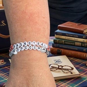 Può includere: Due braccialetti di perline bianche con la scritta "BASKETBALL SOCCER" con accenti di pallone da calcio. È visibile una borsa tote blu navy con "A.C.S." in lettere dorate, insieme a una pila di libri e un paio di occhiali.