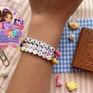 May include: Three beaded bracelets spelling "BRITISH" in white letter beads. The bracelets are stacked on a wrist. Decorative elements include a small brown book, a teapot, and a pink and blue logo with the text "Creative Twinsies".