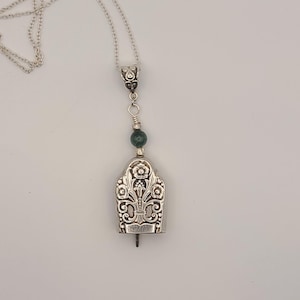 May include: A silver-toned pendant necklace with an ornate, bell-shaped pendant. The pendant features floral and scrollwork designs and is connected to a delicate chain. A green bead accents the design. The necklace is displayed against a white background.
