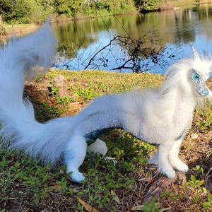 May include: A white, furry dragon with blue eyes and silver scales stands on a grassy bank near a pond. The dragon has a long, flowing tail and a majestic pose.