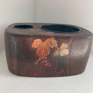 Peut inclure: Un récipient en bois brun foncé avec deux ouvertures circulaires. Le récipient est décoré de feuilles et de baies de couleur cuivrée. Le grain du bois est visible, et le design général est rustique et naturel.