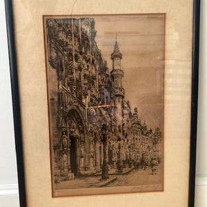 Puede incluir: Un grabado enmarcado que representa una detallada escena callejera europea con arquitectura gótica ornamentada, una torre prominente y un arco a nivel de calle. La obra está en tonos sepia sobre papel, dentro de un marco negro con paspartú beige.