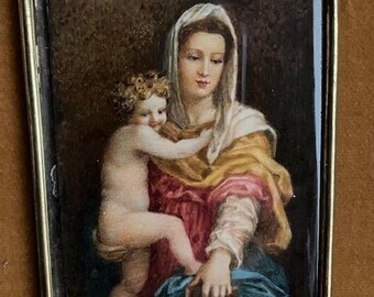 May include: A framed miniature painting depicting a woman and a child. The woman wears a white veil and a red and yellow garment, holding a book. The child is nude. The frame is gold-colored with a small ring at the top for hanging.