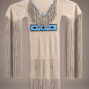May include: A beige suede dress featuring long fringe on the sleeves and hem. The neckline is adorned with a V-shaped fringe collar accented by light blue beads. A rectangular panel across the chest displays a light blue, black, and red pattern.