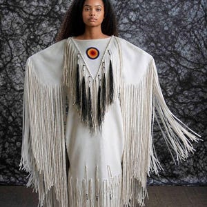 May include: A cream-colored dress with long fringe detailing. The dress features a triangular neckline with a circular design and black fringe accents. The sleeves are long and also have fringe.