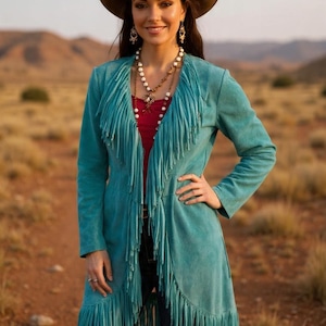 May include: A turquoise suede fringe jacket with a brown cowboy hat and a red top. The jacket has long sleeves and a long fringe detail along the front and hem. The hat has a decorative band.