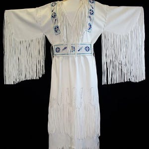 May include: A white dress featuring long sleeves with extensive fringe, a beaded belt, and beaded shoulder panels in blue and white geometric patterns. The garment includes a matching beaded choker and decorative cut-out details along the lower skirt.