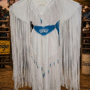 May include: White fringed rodeo outfit with a blue belt and accents. The outfit has long fringe on the sleeves and skirt. The word "RODEO" is visible on a wooden sign in the background.