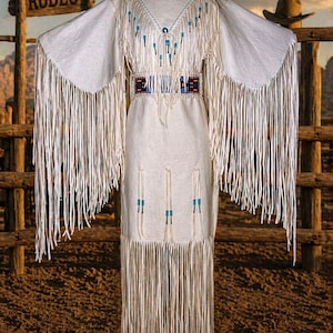 May include: A white dress with long fringe and beaded accents. The dress has a V-neck and a decorative belt. The sleeves extend into fringed panels. The background includes a wooden fence and a sign that says "RODEO".
