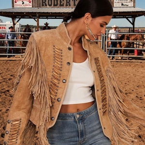 May include: A tan suede jacket with fringe and decorative stitching. The jacket has a stand-up collar, worn with a white top and blue jeans. The background shows a rodeo setting.