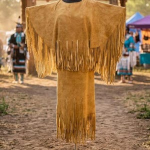 Puede incluir: Un vestido de ante color canela con flecos en las mangas y el dobladillo. El vestido se exhibe en un maniquí, mostrando su diseño y textura. El vestido forma parte de una colección cultural.