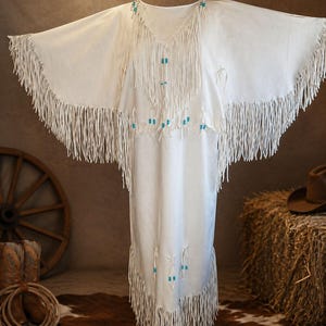 May include: A white, fringed dress with a V-neck and wide sleeves. The dress is adorned with turquoise beads and fringe detailing along the neckline, sleeves, and hem. The dress appears to be made of a soft, flowing fabric.