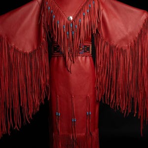 May include: A red leather dress featuring long, flowing fringe along the sleeves and hem. The bodice includes a V-neckline adorned with blue beads and a central silver-toned button. A wide, horizontal band with intricate geometric beadwork sits at the waist.