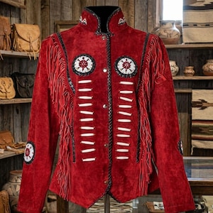 May include: A red suede jacket featuring long fringe along the chest and sleeves, a high collar, and a front button closure. The jacket is decorated with white and black beaded circular patches, white bone-style accents, and braided black trim.
