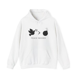Könnte beinhalten: Weißer Kapuzenpullover mit Fronttasche und Kordelzug. Die Grafik zeigt eine schwarze Taube mit Olivenzweig, einen Pfeil und eine Bombe mit dem Text "PEACE NEEDED".