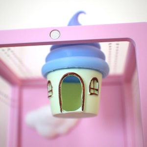 Op de afbeelding: Een grillig miniatuurhuisje met een blauw en wit wervelend dak, een lichtgroen lichaam en bruine raam- en deurkozijnen. Het huisje hangt in een roze, rechthoekig frame, met een witte wolk op de achtergrond. Het thema is fantasie.