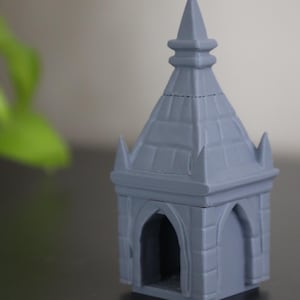 Op de afbeelding: Een klein, grijs, 3D-geprint miniatuurgebouw met een puntig dak en gewelfde deuropeningen. Het model heeft een gedetailleerd, getextureerd oppervlak en een torenspits aan de bovenkant. Het gebouw is een decoratief item.
