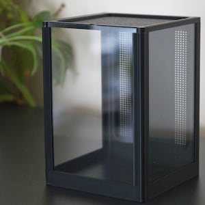 Könnte beinhalten: Ein schwarzes Terrarium mit transparenten Glasscheiben und einer perforierten Seite zur Belüftung. Das rechteckige Gehäuse hat eine Netzoberseite und steht auf einer dunklen Oberfläche. Ideal für die Unterbringung kleiner Reptilien oder Insekten.