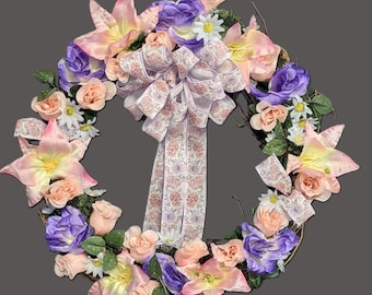 Corona floral primaveral con flores rosas, moradas, lirios, rosas, margaritas y lazo.