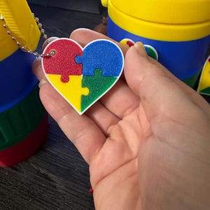 Może przedstawiać: Zawieszka w kształcie serca z czerwonych, niebieskich, zielonych i ż&oacute;łtych element&oacute;w układanki, przymocowana do srebrnego łańcuszka. W tle widoczny jest ż&oacute;łto-niebieski pojemnik.