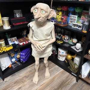 Op de afbeelding: Een levensgrote figuur van Dobby de huiself uit de Harry Potter-serie staat voor een zwarte boekenplank vol thematische merchandise. Dobby draagt een crèmekleurige tuniek. De planken tonen boeken, beeldjes en andere verzamelobjecten.