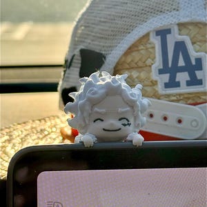 May include: A small white 3D-printed figurine of a character with spiky hair and a smiling expression, positioned on the edge of a black electronic device. Behind the figurine is a straw-textured baseball cap with a blue LA logo on the front.