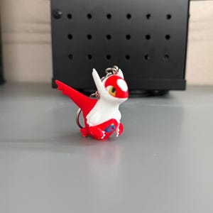 May include: A small, stylized keychain figure of a dragon-like creature with a red and white body. The figure features a prominent red wing, a white head with a red patch, yellow eyes, and a small blue triangular detail on its chest area.