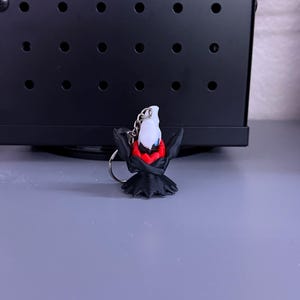 May include: A small, black and red keychain featuring a stylized character with a white head and red accents. The keychain is attached to a silver metal ring. The character has a dark, cape-like design.