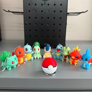 May include: A collection of ten small, colourful plastic character keychains arranged in a semi-circle around a central red and white spherical keychain. The figures include various creatures in shades of green, orange, blue, yellow, and tan.