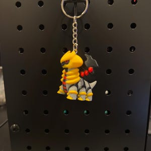 Pode incluir: Um chaveiro com uma criatura amarela, cinza e preta com detalhes vermelhos. A figura está presa a uma corrente e um chaveiro prateados. O fundo é um painel perfurado preto.