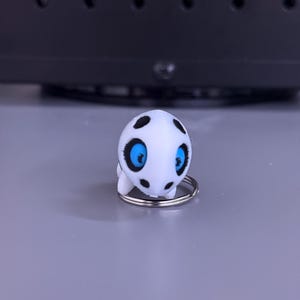 May include: A white, cartoon-like creature keychain with black spots and bright blue eyes. The keychain is attached to a silver metal ring. The creature has a small, rounded body and is standing on a gray surface.