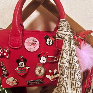 Pode incluir: Uma mala vermelha adornada com v&aacute;rios pins da Minnie Mouse e um len&ccedil;o com padr&atilde;o branco e preto. A mala tem uma pega superior e um fecho de correr. Um pompom rosa pende da pega.