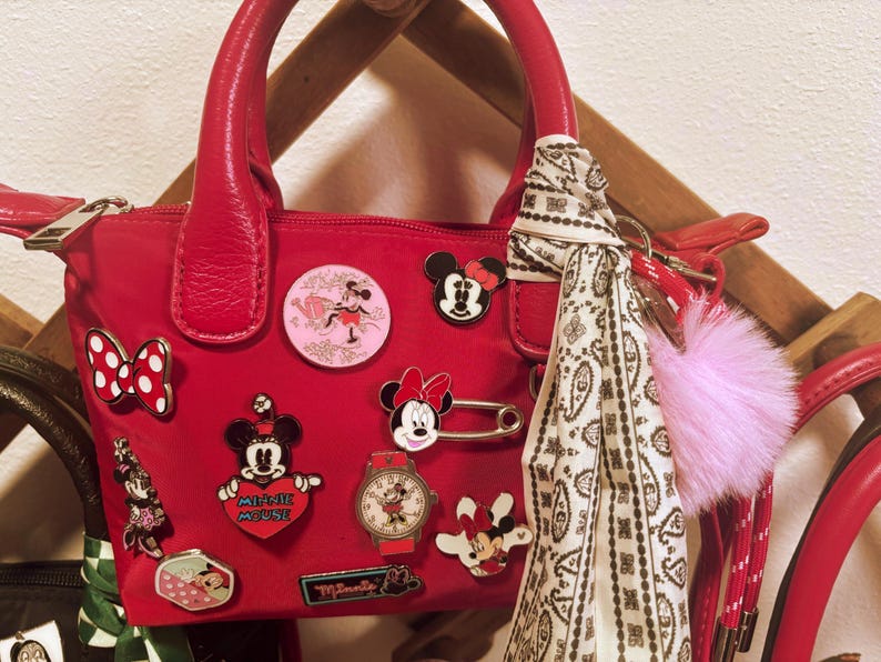 Pode incluir: Uma mala vermelha adornada com v&aacute;rios pins da Minnie Mouse e um len&ccedil;o com padr&atilde;o branco e preto. A mala tem uma pega superior e um fecho de correr. Um pompom rosa pende da pega.