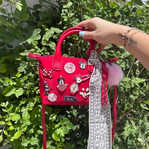 Pode incluir: Uma bolsa vermelha com uma al&ccedil;a superior e uma al&ccedil;a longa. A bolsa &eacute; decorada com v&aacute;rios pinos tem&aacute;ticos da Disney e um len&ccedil;o com estampa branca e preta. Um pompom rosa e borlas vermelhas pendem da bolsa.