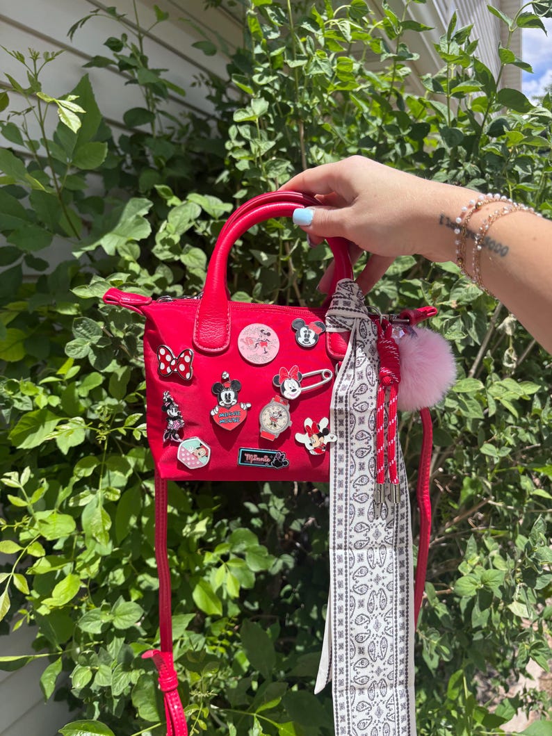 Pode incluir: Uma bolsa vermelha com uma al&ccedil;a superior e uma al&ccedil;a longa. A bolsa &eacute; decorada com v&aacute;rios pinos tem&aacute;ticos da Disney e um len&ccedil;o com estampa branca e preta. Um pompom rosa e borlas vermelhas pendem da bolsa.