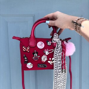 Pode incluir: Uma mala vermelha com uma al&ccedil;a superior e uma al&ccedil;a de ombro remov&iacute;vel. A mala &eacute; decorada com v&aacute;rios pinos tem&aacute;ticos da Disney e um len&ccedil;o paisley branco e preto. Um pompom rosa e um cord&atilde;o vermelho est&atilde;o presos &agrave; mala.