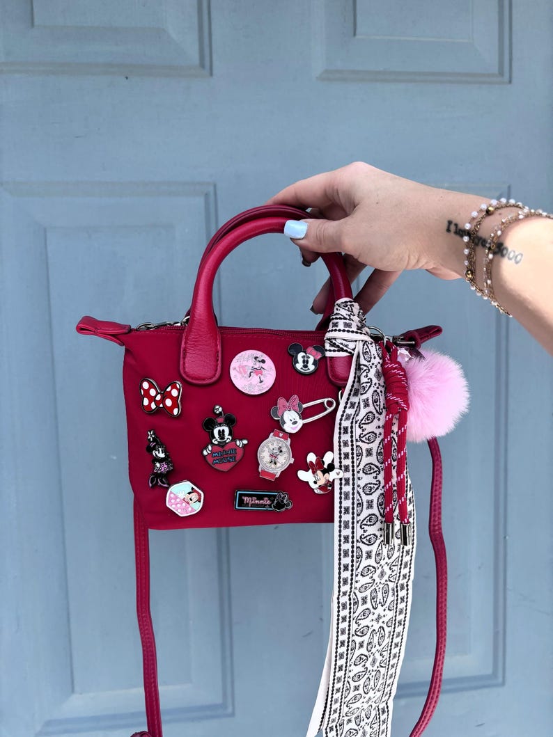 Pode incluir: Uma mala vermelha com uma al&ccedil;a superior e uma al&ccedil;a de ombro remov&iacute;vel. A mala &eacute; decorada com v&aacute;rios pinos tem&aacute;ticos da Disney e um len&ccedil;o paisley branco e preto. Um pompom rosa e um cord&atilde;o vermelho est&atilde;o presos &agrave; mala.