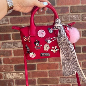 Pode incluir: Uma mala vermelha com uma al&ccedil;a superior e uma al&ccedil;a de ombro remov&iacute;vel. A mala &eacute; decorada com v&aacute;rios pinos tem&aacute;ticos da Disney e um pompom rosa. Um len&ccedil;o estampado est&aacute; amarrado &agrave; al&ccedil;a.