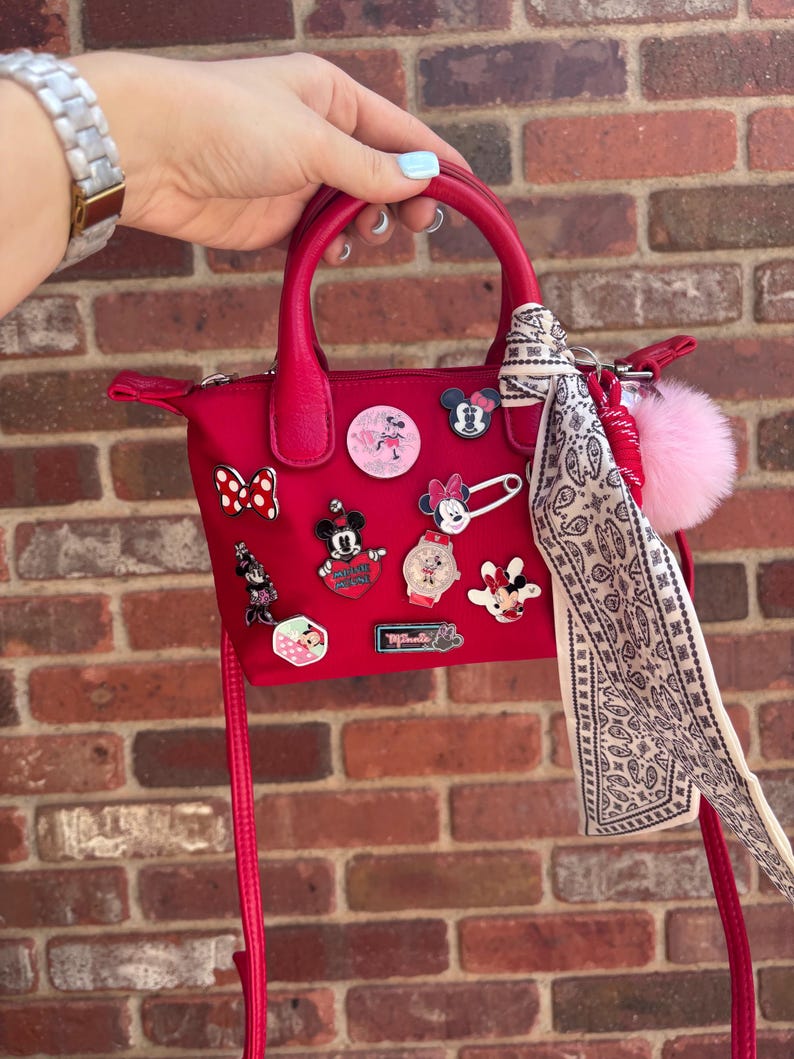 Pode incluir: Uma mala vermelha com uma al&ccedil;a superior e uma al&ccedil;a de ombro remov&iacute;vel. A mala &eacute; decorada com v&aacute;rios pinos tem&aacute;ticos da Disney e um pompom rosa. Um len&ccedil;o estampado est&aacute; amarrado &agrave; al&ccedil;a.
