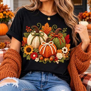 May include: A black t-shirt featuring a graphic print of three crocheted pumpkins in cream, green, and orange. The design includes crocheted sunflowers, red and white flowers, autumn leaves, and acorns arranged around the pumpkins.
