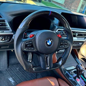 May include: A black and carbon fiber steering wheel with red and blue accents. The wheel has a perforated leather grip and integrated controls. The BMW logo is centered on the airbag cover. The interior features brown leather seats and a digital display.