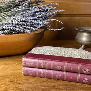 May include: Two antique books with burgundy covers and gold lettering are stacked on a wooden surface. A wooden bowl filled with dried lavender sits behind the books. A small brass pot with three legs is also visible.
